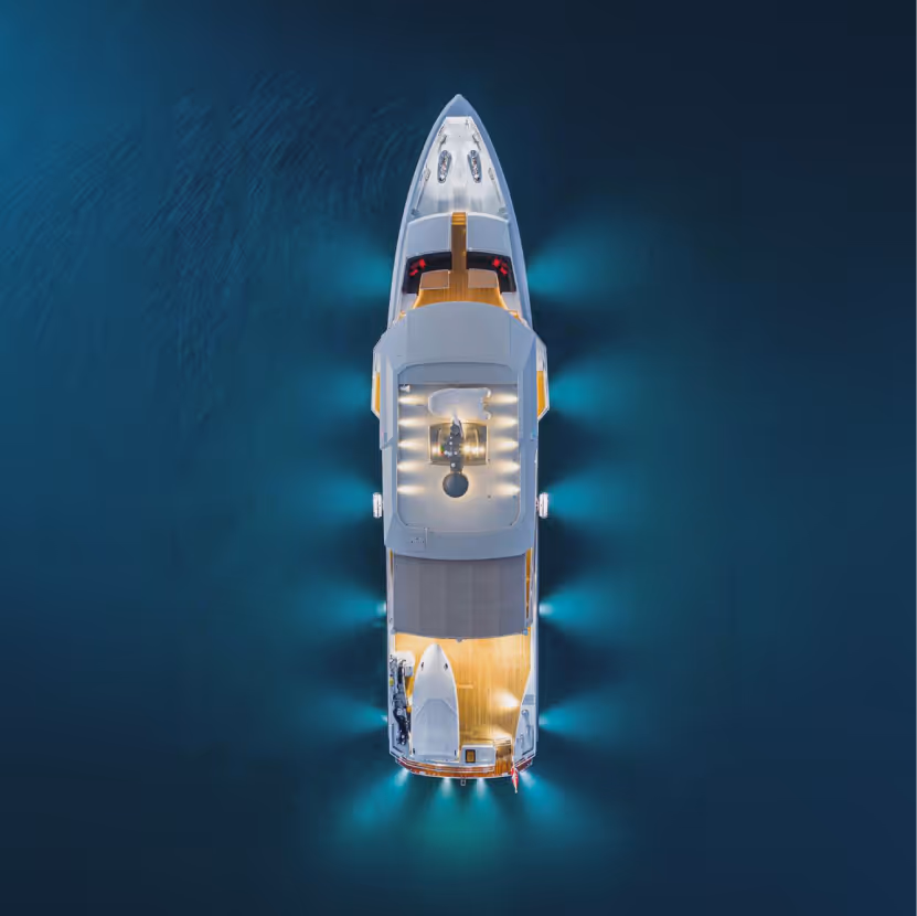 Aerial view of a white yacht illuminated by underwater lights floating on dark blue water at night.