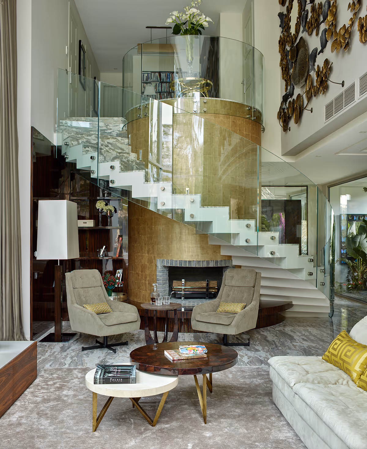 Modern living room with curved glass staircase, two beige armchairs, round coffee tables, and a light sofa with yellow pillows.